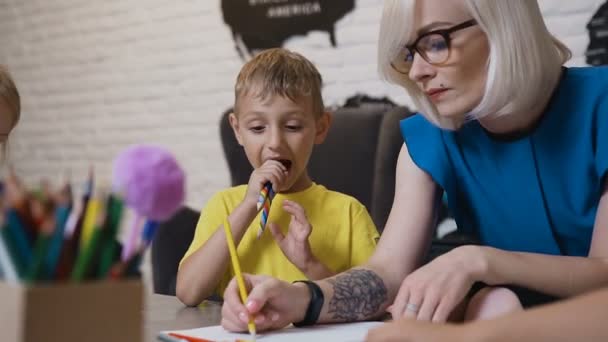 Een jongetje in geel t-shirt met twee kleurpotloden zit aan de tafel en kijken dat zijn leraar tekenen. Tekening leraar en haar kleine student — Stockvideo