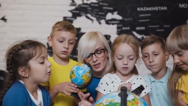 Zbliżenie, chłopców, dziewcząt i ich nauczyciel youn, którzy studiują na mapie świata za pomocą kuli ziemskiej i szkło powiększające. Atrakcyjne nauczyciel naucza pięć z jego uczniów szkoły podstawowej — Wideo stockowe