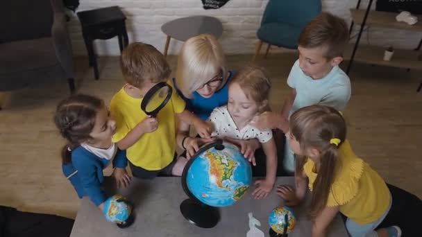 Вид сверху. Маленькие кавказские ученики мальчиков и девочек смотрят на глобусы, слушая учителя на уроке географии. Один из мальчиков смотрит на земной шар через лупу — стоковое видео