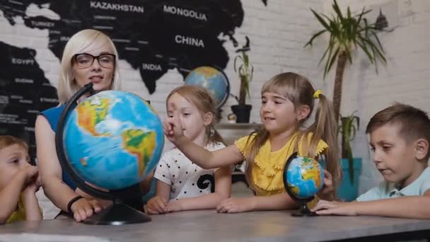 Belos alunos e professores olham para globos em sala de aula de geografia sobre a lição na escola primária. Meninos, meninas e seu jovem professor estudar o mapa do mundo usando o globo e — Vídeo de Stock