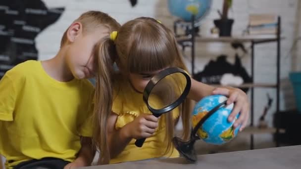 Une belle fille avec un garçon à travers la loupe regarde le globe dans la salle de classe. Lors d'une leçon de géographie, les enfants étudient le globe. Voyager autour du monde — Video