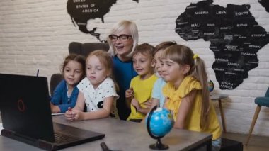 Bilgisayar terminali ilköğretim okulunda beyaz çocuklara yardımcı öğretmen. Coğrafya dersinde öğretmen ile bilgisayar kullanarak gülümseyen okul çocukları. Şık kadın ve İlköğretim öğrencilerinin dizüstü bilgisayar kullanarak