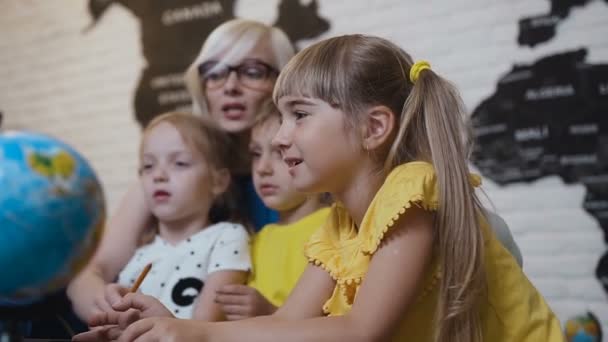 Un niño lindo con el profesor se sienta a la mesa en el aula de geografía y está curiosamente estudiando el globo terráqueo o el mapa del mundo. Pequeños alumnos miran el globo en el aula — Vídeo de stock