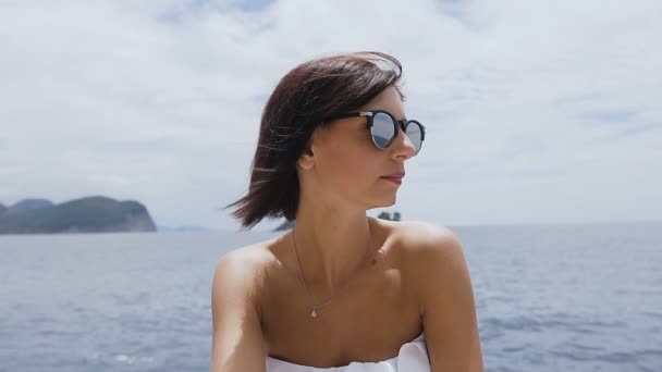 Красивая девушка сидит на скале в солнечных очках, в белом купальнике на фоне морской голубой воды. Адриатическое море. Летний отдых — стоковое видео