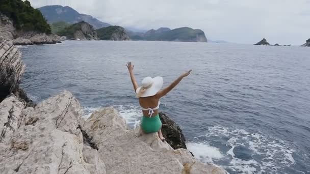 Mujer joven vestida con traje de baño y sombrero se sienta en el borde de los acantilados levantando las manos y mirando hacia un mar azul adriático Concepto de vacaciones, aventura y estilo de vida — Vídeos de Stock