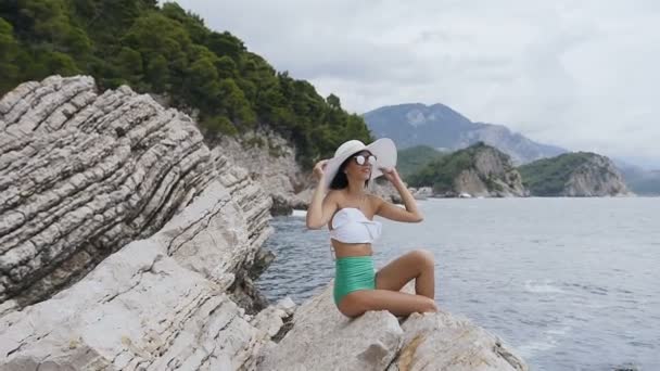 Joven mujer bonita en gafas de sol en traje de baño y con sombrero ancho, se sienta en las rocas cerca del mar. Vacaciones de verano. Mar Adriático, Montenegro — Vídeos de Stock