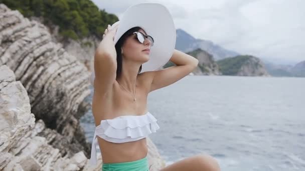 Mujer joven en sombrero y traje de baño sentada en una roca y mira el mar Adriático en Petrovac, Montenegro. Chica atractiva relajándose cerca del mar. Vacaciones de verano — Vídeo de stock