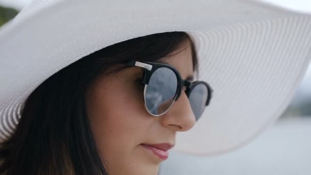 Portrait of a young woman with big hat and sunglasses enjoying sun and water. A pretty young brunette woman with big elegant hat and sunglasses, enjoy sunny day near blue ocean, vacation time. Close — Stock Video