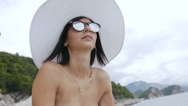 Junge Frau im weißen Badeanzug mit großem Hut und Sonnenbrille, die Sonne und Wasser am Felsenstrand genießt. Sommerurlaub am Meer — Stockvideo