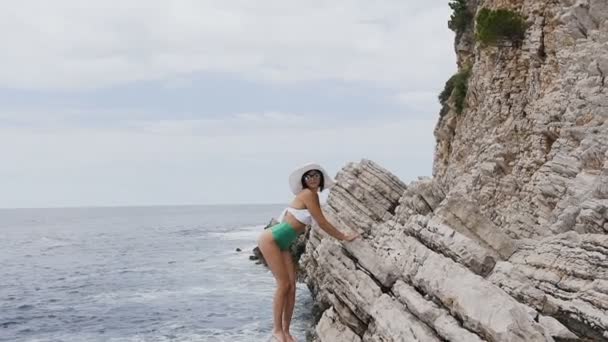 Cute slim tanned girl in a stylish swimsuit and a big hat poses on the camera, standing by the rocky cliff against the background of the sea. Summer vacation on the sea — Stock Video
