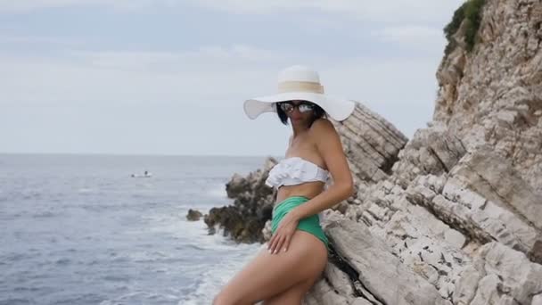 Carino snello abbronzato ragazza in un elegante costume da bagno e un grande cappello posa sulla macchina fotografica, in piedi vicino alla scogliera rocciosa sullo sfondo del mare. Sexy ragazza attraente vicino roccia sullo sfondo mare — Video Stock