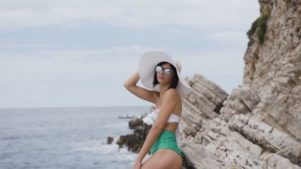 Sexy morena delgada vestida de blanco con bikini verde, gafas de sol y sombrero grande, posando en la playa cerca de la roca en un día soleado sobre el fondo del mar. Hermosa chica con piernas delgadas en la playa cerca — Vídeo de stock