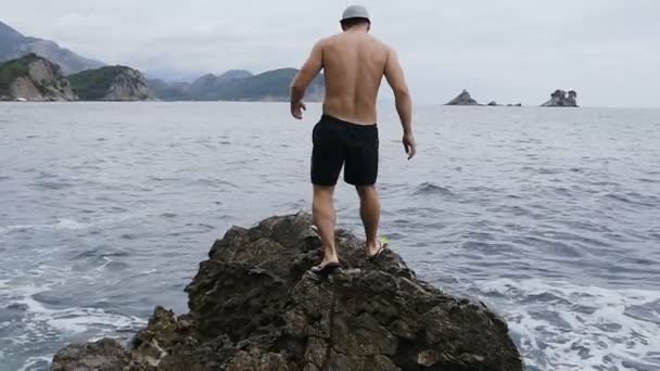 Fröhlicher junger Mann hebt seine Hände hoch oben auf der Klippe über dem Meer bei schönen Wellen Meer und Felsen Hintergrund. Sommerurlaub auf dem Meer. Urlaub in Montenegro — Stockvideo