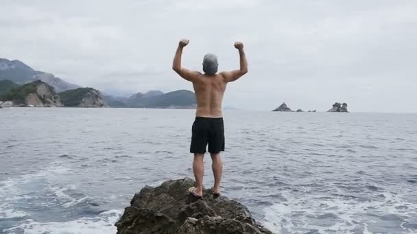 Молодой человек поднимает руки к небу, стоя на камнях у моря. Веселый человек поднимает руки высоко на вершине скалы над морем, где прекрасный вид на волны и скалы — стоковое видео