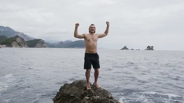 Una persona se regocija en la vida levantando sus manos sobre una roca junto al mar. Vacaciones en el mar Adriático. Viaje de verano — Vídeos de Stock