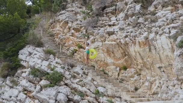 Chica delgada vestida con traje de baño blanco-verde y sombrero grande está caminando sobre pasos cerca de rocas altas a la playa con anillo inflable. Mujer joven con anillo flotante saludando su mano y va por la — Vídeos de Stock