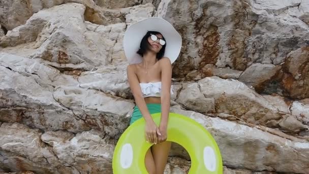 Mujer joven bronceada en bikini blanco, gafas de sol y sombrero grande con colchón cerca de la roca alta junto al mar. Vacaciones de verano. Día de verano — Vídeo de stock