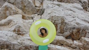 Çekici kız bir mayo ve bir şapka yapar elinde bir şişme float daire komik bir cilvesi olduğunu ve kaya arka plan kameraya bakar. Sarı şişme float tabaklanmış kadınla