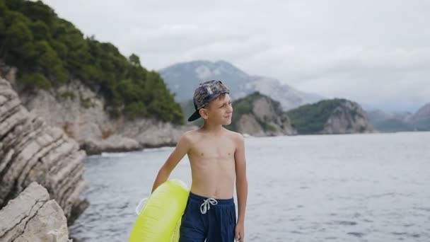 Attraktiver Junge beobachtet Meer und steht auf einer Klippe mit aufblasbarem Kreis. Ein kleiner Junge steht mit einem gelben Schlauchboot auf dem Felsen und genießt die Schönheit der Landschaft. niedlichen Jungen 7-8 Jahre liebt — Stockvideo