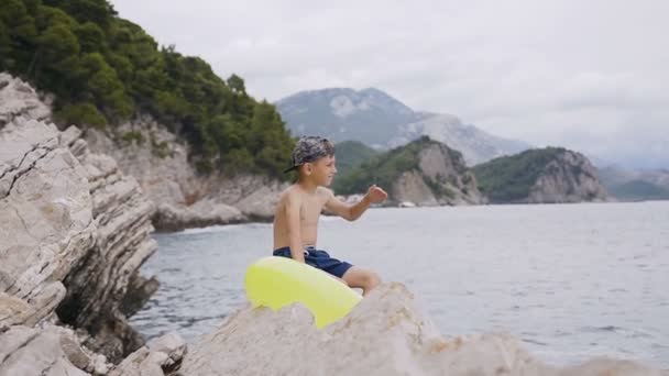 Επτά-οκτώ χρόνια αγόρι κάθεται σε βράχο και βλέπουν ένα όμορφο ορίζοντα. Χαριτωμένο αγόρι με φουσκωτό δαχτυλίδι που κάθεται σε βράχο κοντά του δίπλα στη θάλασσα. Καλοκαιρινές διακοπές — Αρχείο Βίντεο