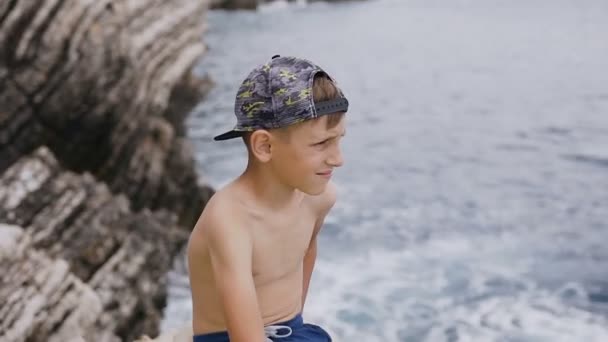 Ein attraktiver Typ mit Mütze sitzt auf einem Felsen vor dem Hintergrund des Meeres und der Felsen zur Sommerzeit oder im Urlaub. Netter Junge in Mütze und kurzer Hose sitzt auf einem Felsen und blickt auf das blaue Meer. Attraktiv wenig — Stockvideo