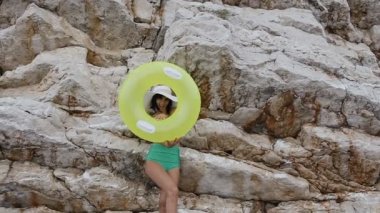 İnce güzel esmer yeşil mayo ve şişme sarı float veya kaya arka plan daire kameradan büyük şapka gülümsüyor. Şişme float tabaklanmış genç kadın