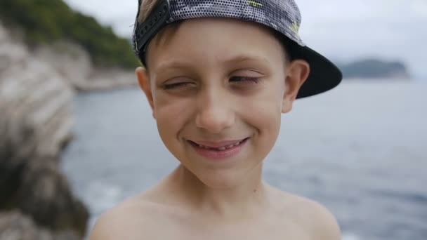 Ελκυστικό μικρό αγόρι με τα μπλε μάτια βλέπουν φωτογραφική μηχανή. Γκρο πλαν, από ένα μικρό αγόρι στο καπάκι να αναβοσβήνει τα μάτια σχετικά με την κάμερα στο παρασκήνιο στη θάλασσα. Καλοκαιρινές διακοπές — Αρχείο Βίντεο