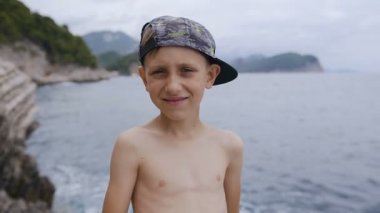 Kayanın üzerinde kameraya gülümseyen ve gösterilen süre ayakta beyaz küçük çocuk denizde arka plan yaşasın. Yaz tatili, tatil çocuk. Sivri şapkalı hoş gülümseyen çocuk portresi