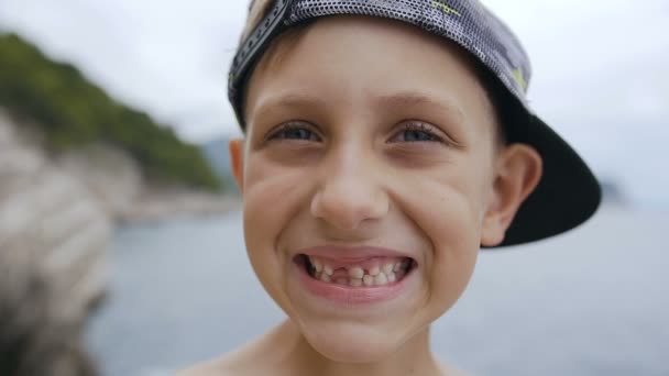 Un ragazzo senza denti carino in berretto sorride sulla macchina fotografica sullo sfondo del mare. Ritratto di uno scolaro sorridente senza denti — Video Stock