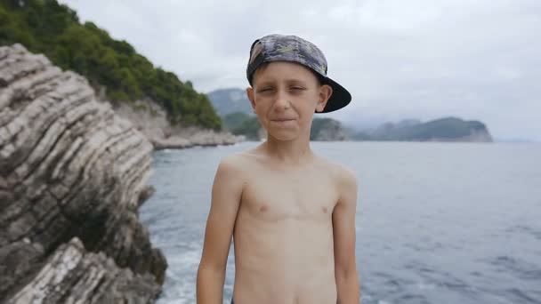 白人小男孩站在岩石上, 微笑着看着镜头, 在海面上露出大拇指。暑假, 孩子在度假。一个可爱的微笑男孩在尖顶帽子的肖像 — 图库视频影像