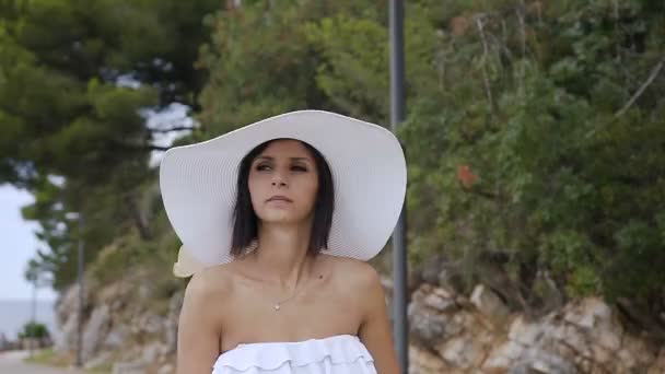 Menina morena atraente em um chapéu grande desce no parque perto da praia. Retrato de uma jovem mulher em um chapéu que caminha perto do mar ao ar livre — Vídeo de Stock