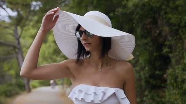 Porträt einer hübschen jungen Frau, die an einem sonnigen, warmen Sommertag im Park am Meer weißen Badeanzug und großen Hut trägt. Spaziergänge im Sommerpark — Stockvideo