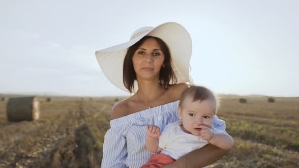 Portrait of a beautiful young woman in a dress with a big hat and her little son in a white body standing in a golden field with bales in the sunset. Warm autumn, harvest, field, bales of straw — Stock Video