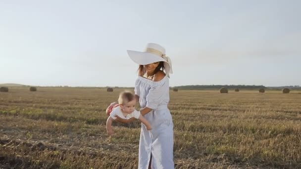 Atractiva mujer joven en vestido y sombrero se divierte con su bebé en el campo al atardecer. Mamá e hijo están jugando en el campo con fardos del día de verano — Vídeos de Stock
