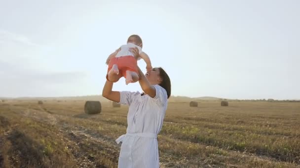 Piękna kobieta wysoko podnosi jej adorable baby boy i zaczyna, odwracając ją uśmiechając się. Szczęśliwa młoda kobieta spędzać czas, grając z synem w polu o zachodzie słońca — Wideo stockowe