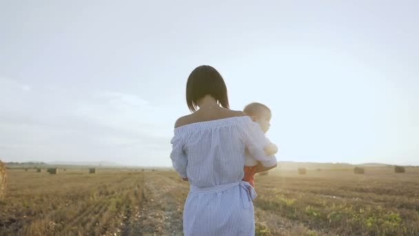 Vista traseira. Uma jovem mãe carrega um pequeno filho em seus braços andando no campo ao pôr-do-sol. Caminhe ao pôr do sol menina e pequeno filho no campo com fardos de palha no verão ou no outono — Vídeo de Stock