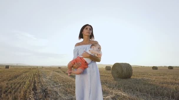Attraente giovane donna porta piccolo figlio in giro per il campo tra le braccia al tramonto. Mamma e bambino in una passeggiata serale — Video Stock