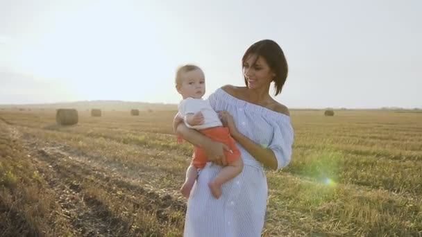 Porträtt av glad kaukasiska mor som innehar hennes lille son, hon leker med barnet i fältet vid solnedgången. Glad ung kvinna leker med lilla son i fältet med balar vid solnedgången — Stockvideo