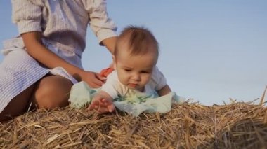C Genç Anne mavi elbiseli bir haystack yaz oynarken beyaz gömlekli güzel bir çocuk. Saman balya üzerinde yatan bir bebek tutan oynayan kadın