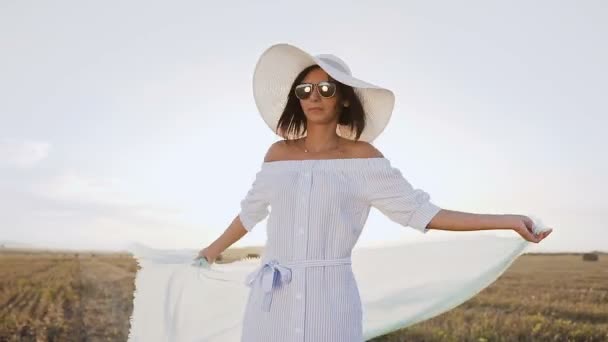 Porträtt av en attraktiv tjej i en lång klänning, solglasögon, stor hatt med en halsduk snurrar runt själv i fältet vid solnedgång sommar. Ung kvinna gå på gyllene fält. Frihet-konceptet. Lycklig kvinna — Stockvideo