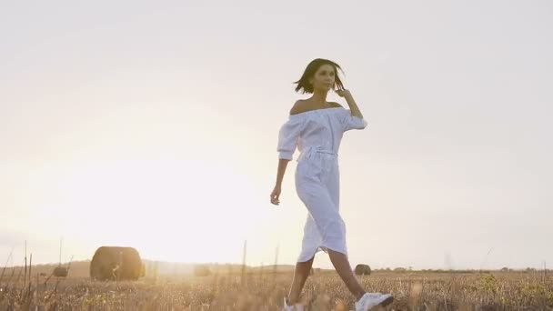 Uma jovem correndo no campo faz círculos em torno dele em uma noite de verão ao pôr-do-sol. Menina atraente em um vestido longo vai um rastro no campo com fardos de palha — Vídeo de Stock