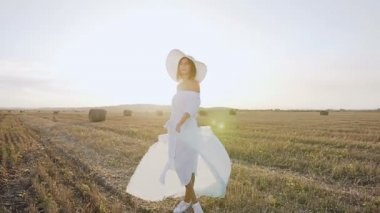 Uzun elbiseli çekici kız, bir eşarp ile büyük şapka günbatımı yaz alanında kendini döner. Sarı saman balya sahada yürüyen genç kadın güzellik. Özgürlük kavramı. Mutlu kadın açık havada