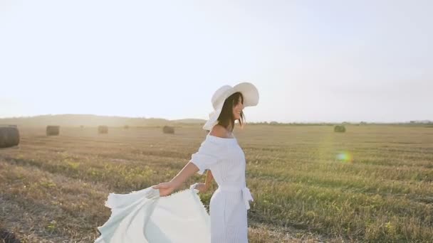 Привлекательная улыбающаяся девушка в длинном платье, большая шляпа с шарфом крутится вокруг себя в поле на закате лета. Красавица, молодая женщина ходит по полю из желтых соломенных тюков. Понятие свободы. Счастливая женщина — стоковое видео