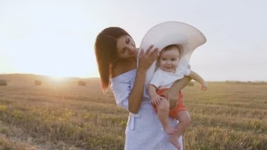 Büyük bir şapka alanında annesiyle günbatımında yürüyüş bir bebek portresi. Mutlu genç kadın alanında yaz akşamları oynarken küçük oğlu eğlenceli ile