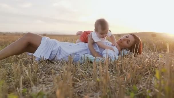 Junge Frau mit ihrem entzückenden 6 Monate alten Jungen, der draußen auf dem Feld spielt. eine junge Mutter mit einem kleinen Sohn auf dem Arm liegt im Sommer im Sonnenuntergang auf dem Feld — Stockvideo