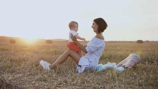 En ung kvinna i en ljus klänning sitter på gräset i fältet och leker med en underbar bebis. Lycklig mamma med son promenader i fältet i sommarkväll. Halmbalar i fältet. — Stockvideo