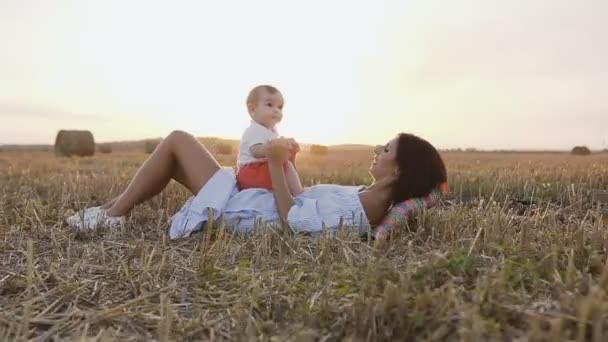 Jovem mãe está segurando seu filho em suas mãos brincando de diversão com ele no campo no verão ao pôr do sol. Cuidados maternos e amor para o menino na natureza — Vídeo de Stock