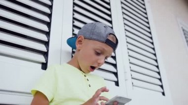 Yüzü ve elleri 5-6 yıl yaşlı çocuk yakınındaki ayakta bilgisayar oyunları oynarken açık pencerede beyaz kapatın. Telefon kullanarak evlat. Teen ve teknoloji kavramı