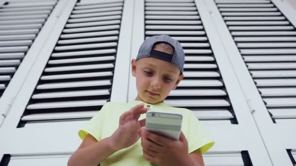 Primer plano del niño pequeño usando el teléfono móvil mientras juega. Retrato de un lindo chico con camiseta escribiendo un mensaje usando un teléfono inteligente, mirando y sonriendo en la pantalla, disfrutando de las vacaciones de verano — Vídeo de stock