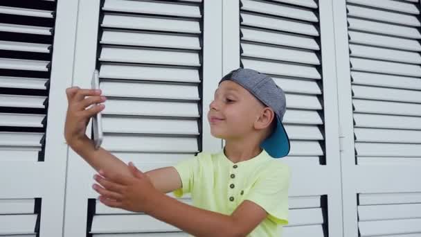 Lindo niño pequeño en camiseta y gorra haciendo foto selfie con teléfono inteligente en casa aislado sobre fondo blanco. Feliz adolescente haciendo foto selfie para las redes sociales — Vídeo de stock